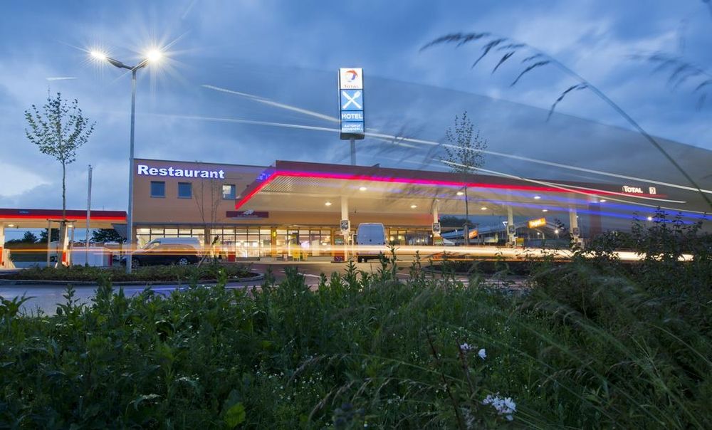Tankstelle mit Restaurant in der Abenddämmerung, Neonlichter eingeschaltet, Langzeitbelichtung mit Lichtspuren von Autos.